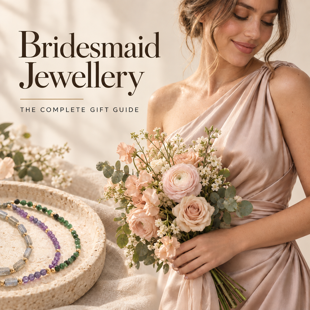 Bridesmaid Jewellery, the complete gift guide — a bridesmaid in a blush satin dress holding a pastel rose bouquet, with labradorite, amethyst and green gemstone bracelets on a stone dish