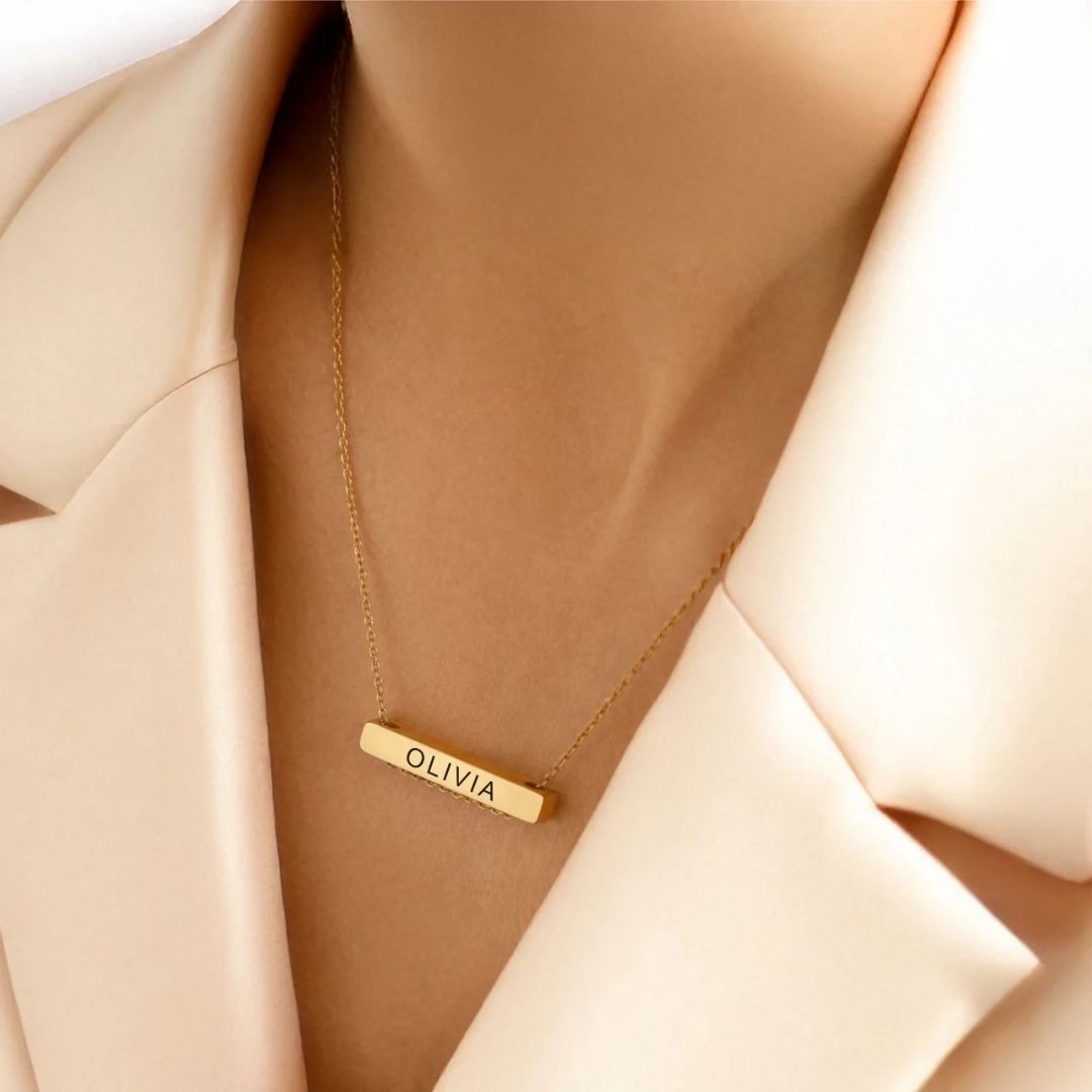 A model in a cream blazer wearing a waterproof 18K gold plated custom engraved bar name necklace, showcasing a single personalised stainless steel pendant.
