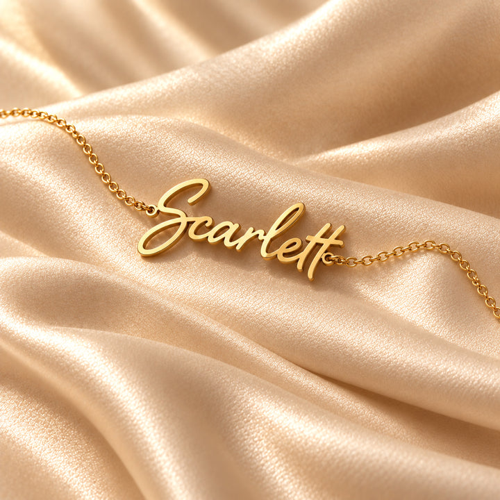 Gold script name bracelet on a dainty chain, flat lay on satin