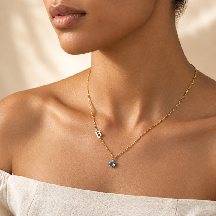 Model wearing an 18K gold sideways initial necklace with letter B and a blue March birthstone charm