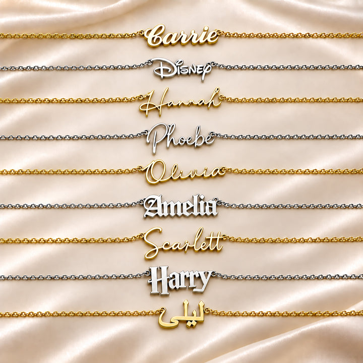 Nine personalised name necklaces are arranged horizontally on a draped champagne satin background. The necklaces feature custom script names including Carrie, Hannah, Phoebe, Olivia, Amelia, Scarlett, Harry shown in both gold and silver metal finishes.