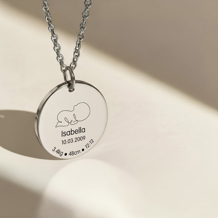 Silver new mum necklace with a round pendant engraved with baby birth details on a beige background