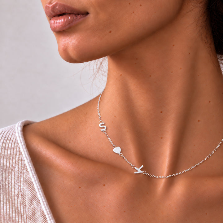 Silver sideways initial necklace worn on a model — letters S and K with a heart charm between them