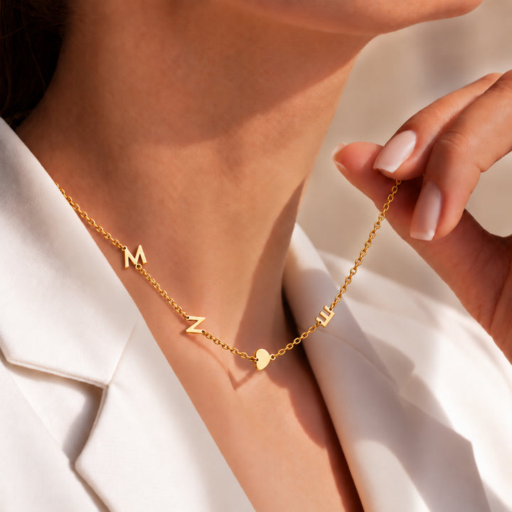 Gold sideways initial necklace with three initials and a heart charm, worn with an open collar