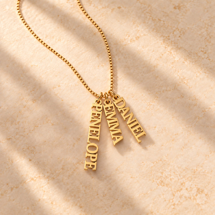 Gold vertical name plate necklace with three names in block capitals, flat lay